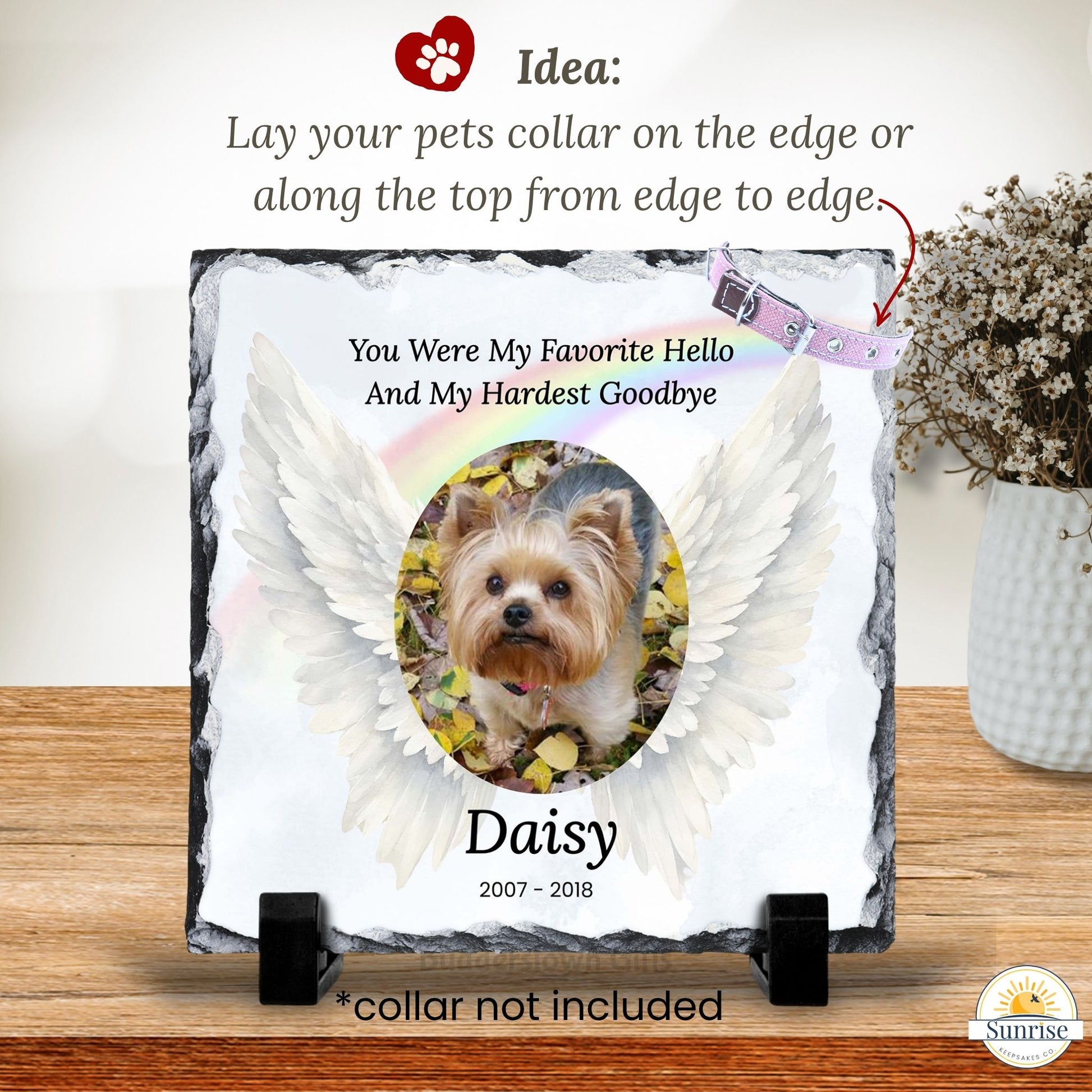 Personalized pet memorial slate plaque on a wooden surface, featuring a photo of a Yorkshire Terrier between white angel wings with the text 'You Were My Favorite Hello And My Hardest Goodbye' and the name 'Daisy 2007 - 2018'. A pink pet collar is shown at the top right with an arrow and text explaining the layout, and a small 'Sunrise' logo appears in the bottom right corner.