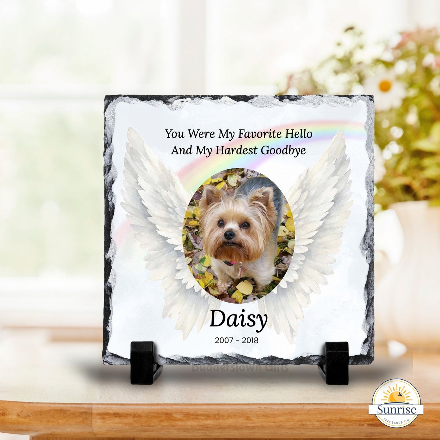 Personalized pet memorial slate plaque on a small black stand, featuring a photo of a Yorkshire Terrier with white angel wings and the text 'You Were My Favorite Hello And My Hardest Goodbye Daisy 2007-2018'. The plaque is displayed on a wooden surface with a blurred indoor background.