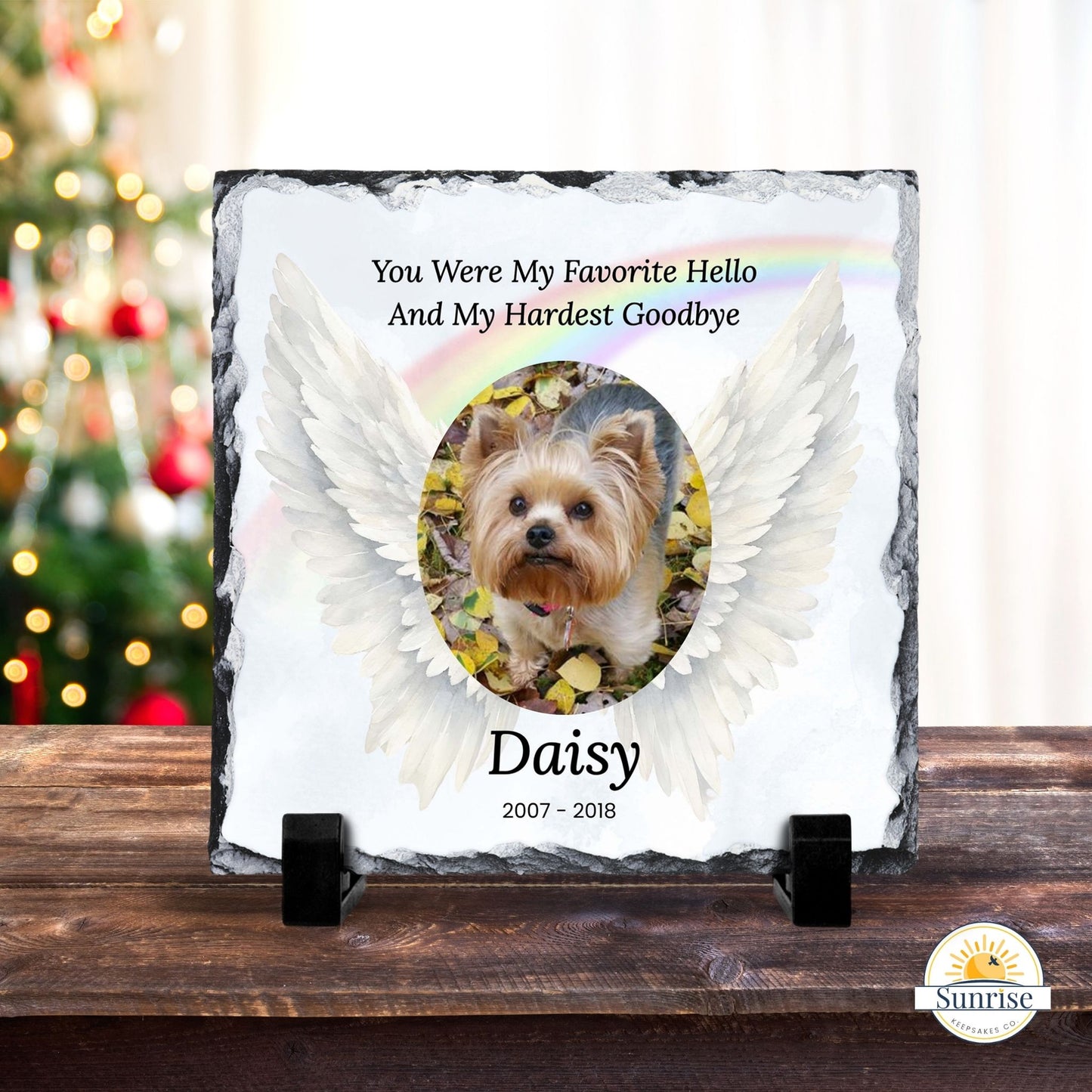 Personalized memorial slate plaque on a wooden stand, featuring a photo of a Yorkshire Terrier surrounded by white angel wings and the text 'You Were My Favorite Hello And My Hardest Goodbye' and 'Daisy 2007 - 2018'. A blurred Christmas tree and warm indoor background are visible behind it.
