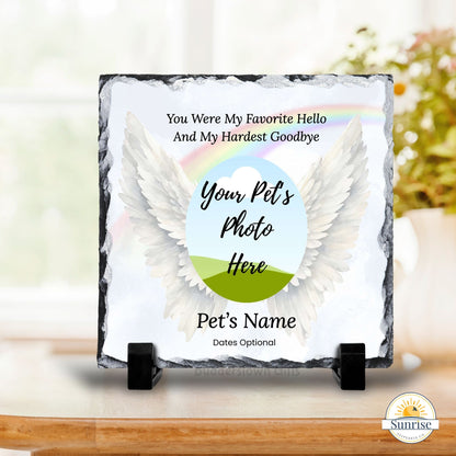 Personalized pet memorial slate plaque on a small black stand, featuring white angel wings, a pastel rainbow, and customizable text including a pet photo circle and the name 'Pet's Name'. Shown on a tabletop with a softly blurred indoor background.