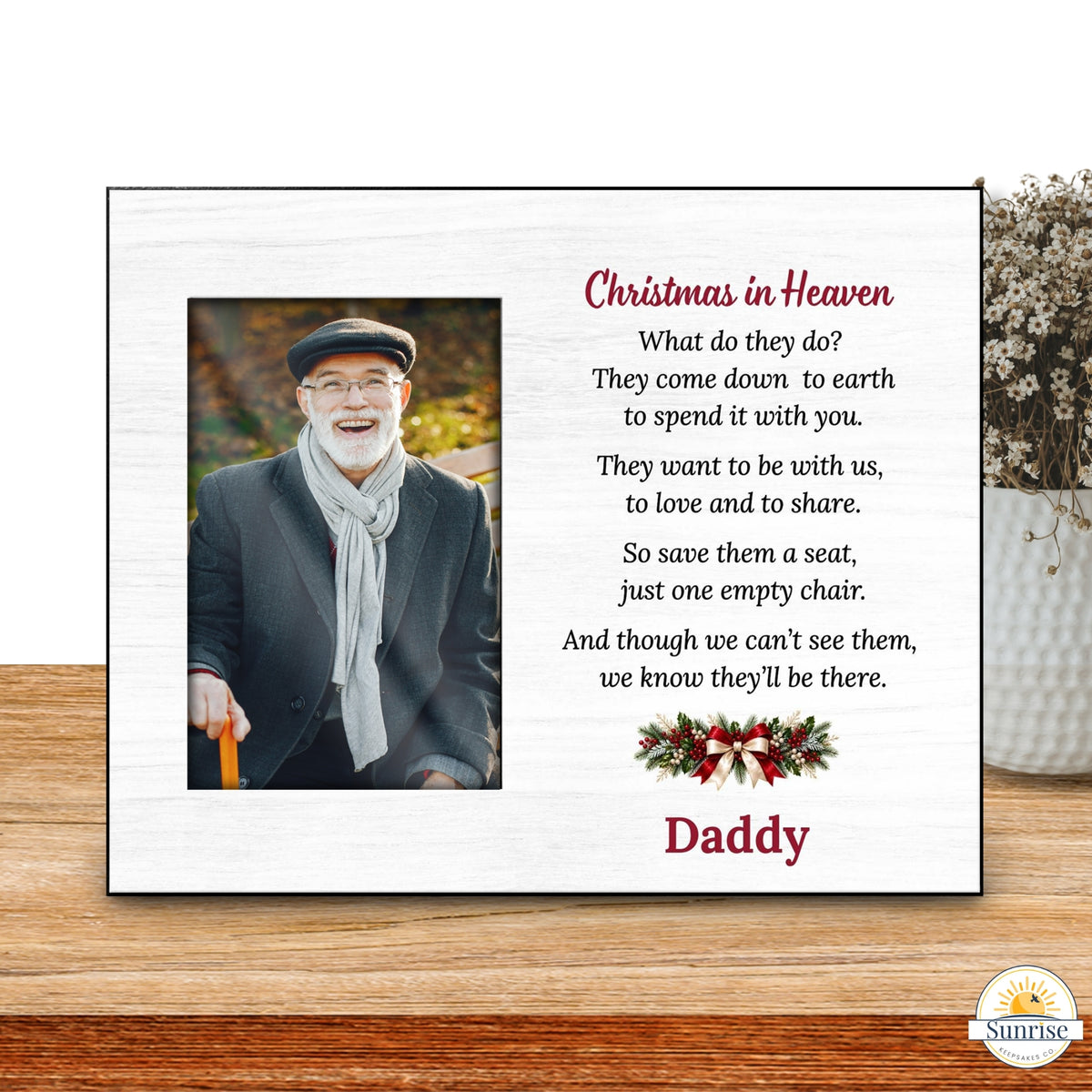 A personalized Christmas in Heaven memorial plaque displayed on a wooden tabletop next to a vase of dried flowers. The white plaque features a photo of a smiling man, a poem about the afterlife, and the name 'Daddy' in red script at the bottom.