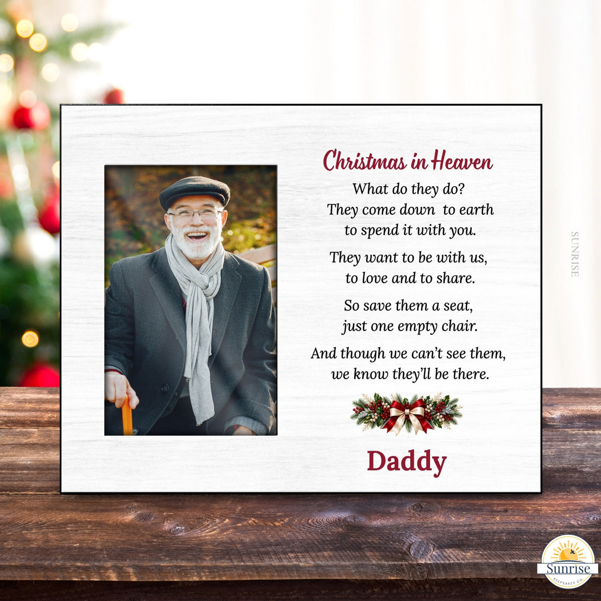A personalized Christmas in Heaven memorial plaque displayed on a wooden surface with a blurred Christmas tree in the background. The white plaque features a photo of a smiling man, a poem, a small holiday wreath, and the name 'Daddy' in red text.