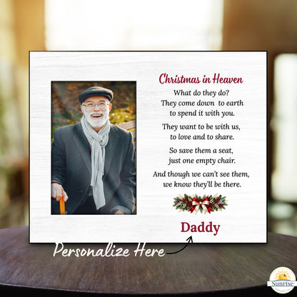 A personalized Christmas in Heaven memorial plaque displayed on a dark wooden table with a blurred indoor background. The white plaque features a photo of a smiling man, a poem, a red 'Daddy' name, and a holiday wreath.