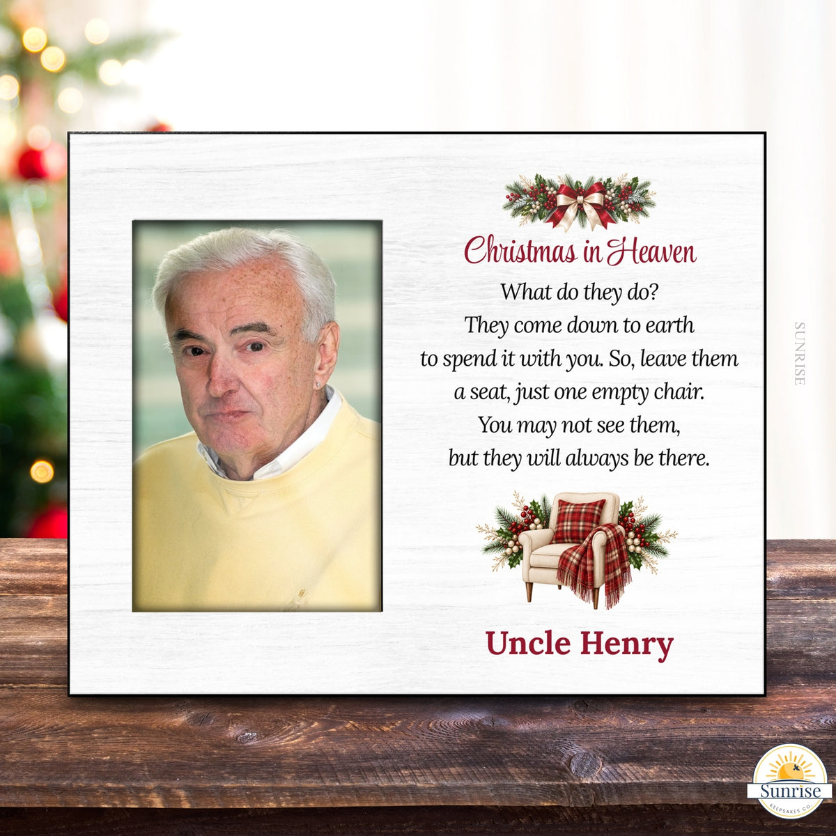 A personalized Christmas in Heaven memorial plaque shown on a wooden tabletop with a blurred Christmas tree in the background. The white plaque features a photo slot for a portrait, red and black text, holiday greenery and red plaid accents, and the name 'Uncle Henry'.