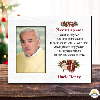A personalized Christmas in Heaven memorial plaque shown on a wooden tabletop with a blurred Christmas tree in the background. The white plaque features a photo slot for a portrait, red and black text, holiday greenery and red plaid accents, and the name 'Uncle Henry'.