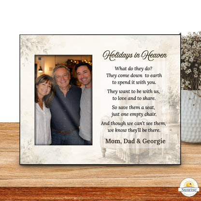 A personalized framed memorial sign displayed on a wooden tabletop. The sign has a cream background with floral corner details, a photo of three people on the left, and a poem titled 'Holidays in Heaven' on the right, personalized with the names 'Mom, Dad & Georgie'.