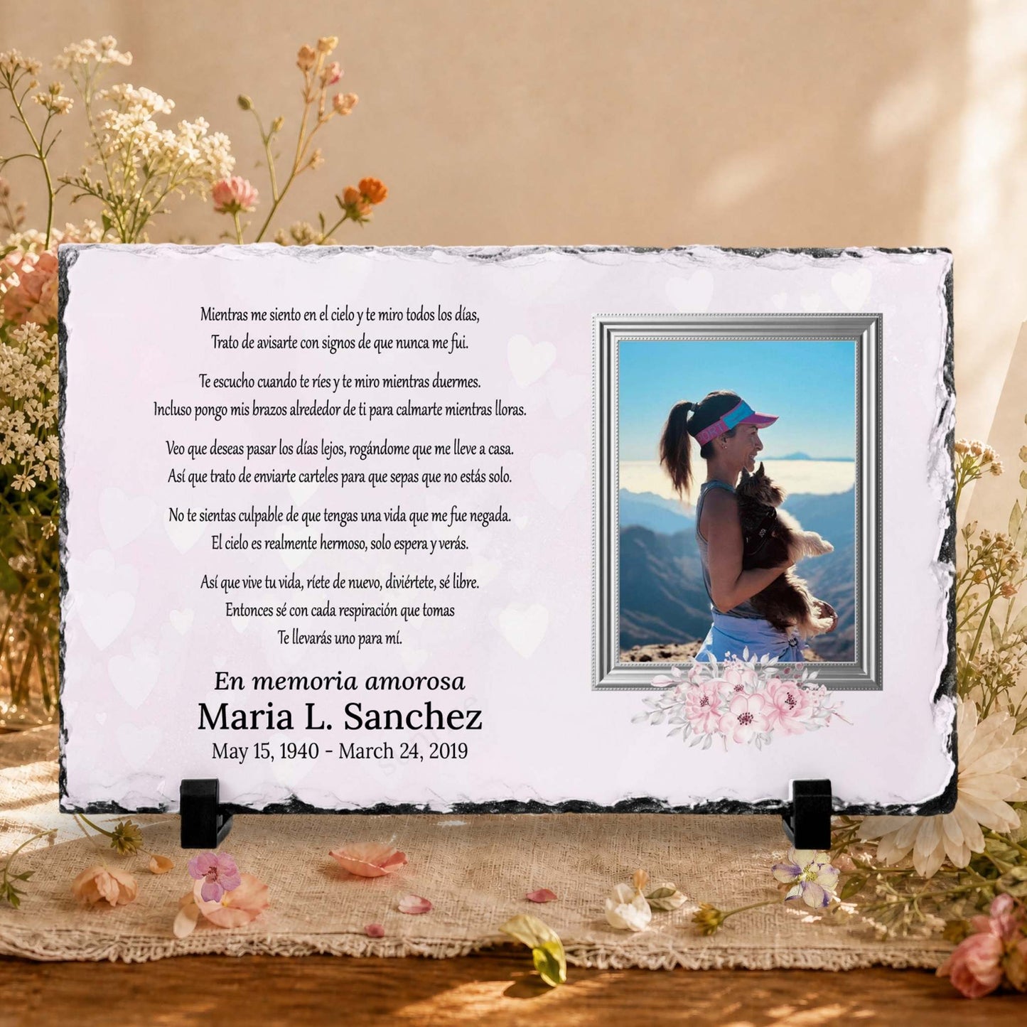 A memorial stone plaque with Spanish text, a photo of a woman and the names and dates of loved one .  memorial slate displayed on a burlap surface with dried flowers.
