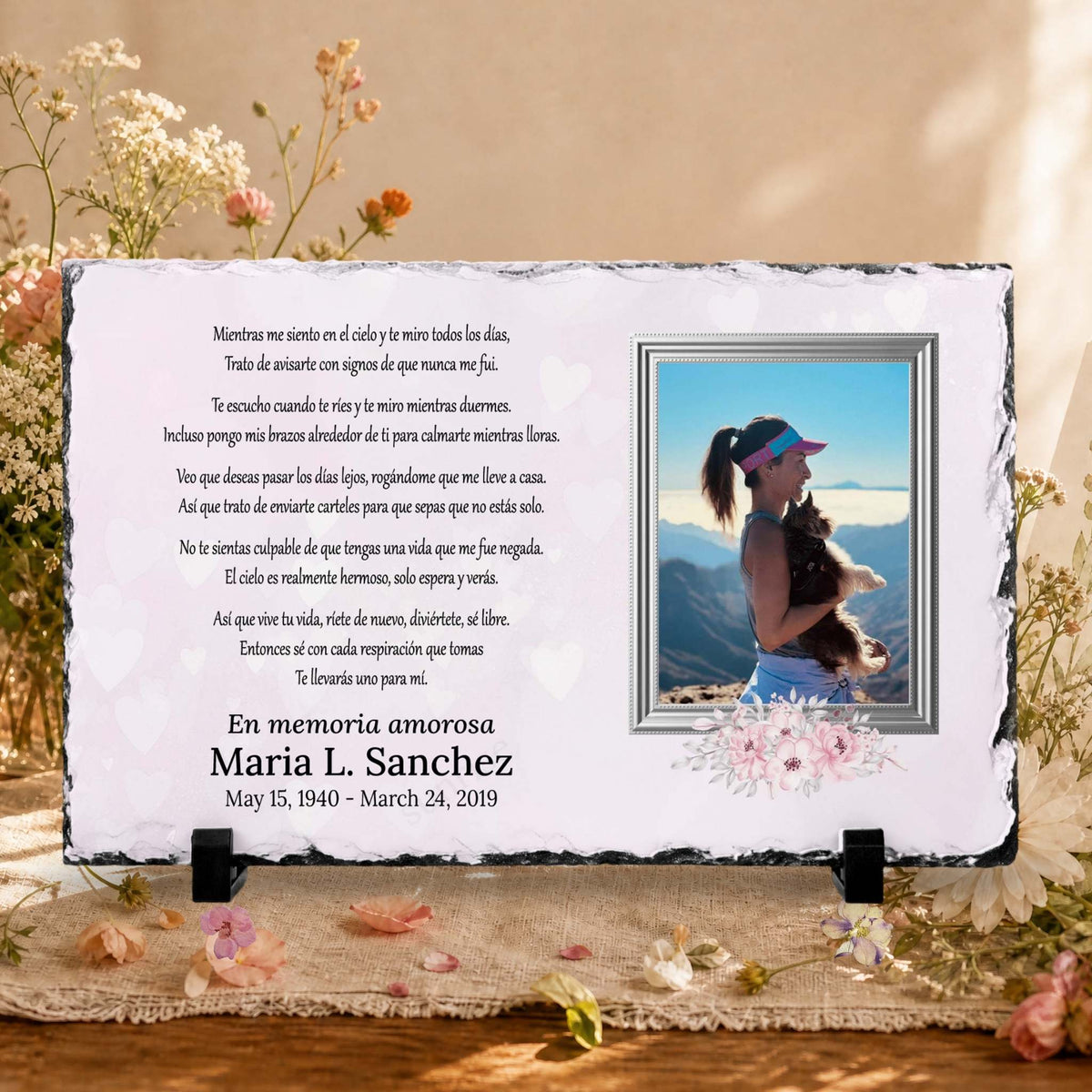 A memorial stone plaque with Spanish text, a photo of a woman and the names and dates of loved one .  memorial slate displayed on a burlap surface with dried flowers.