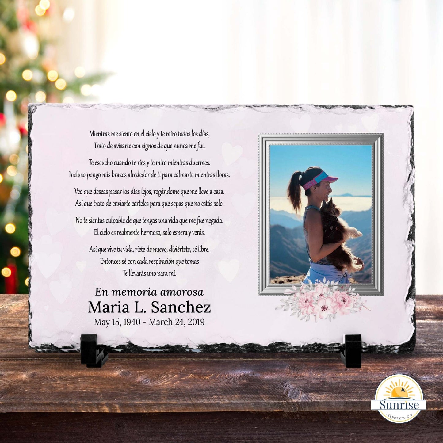 Personalized memorial stone plaque on a wooden surface, featuring a Spanish remembrance poem, the names and dates of a deceased loved one, and a small inset photo of a woman. The plaque is displayed on two small black stands against a blurred festive background with a Christmas tree.