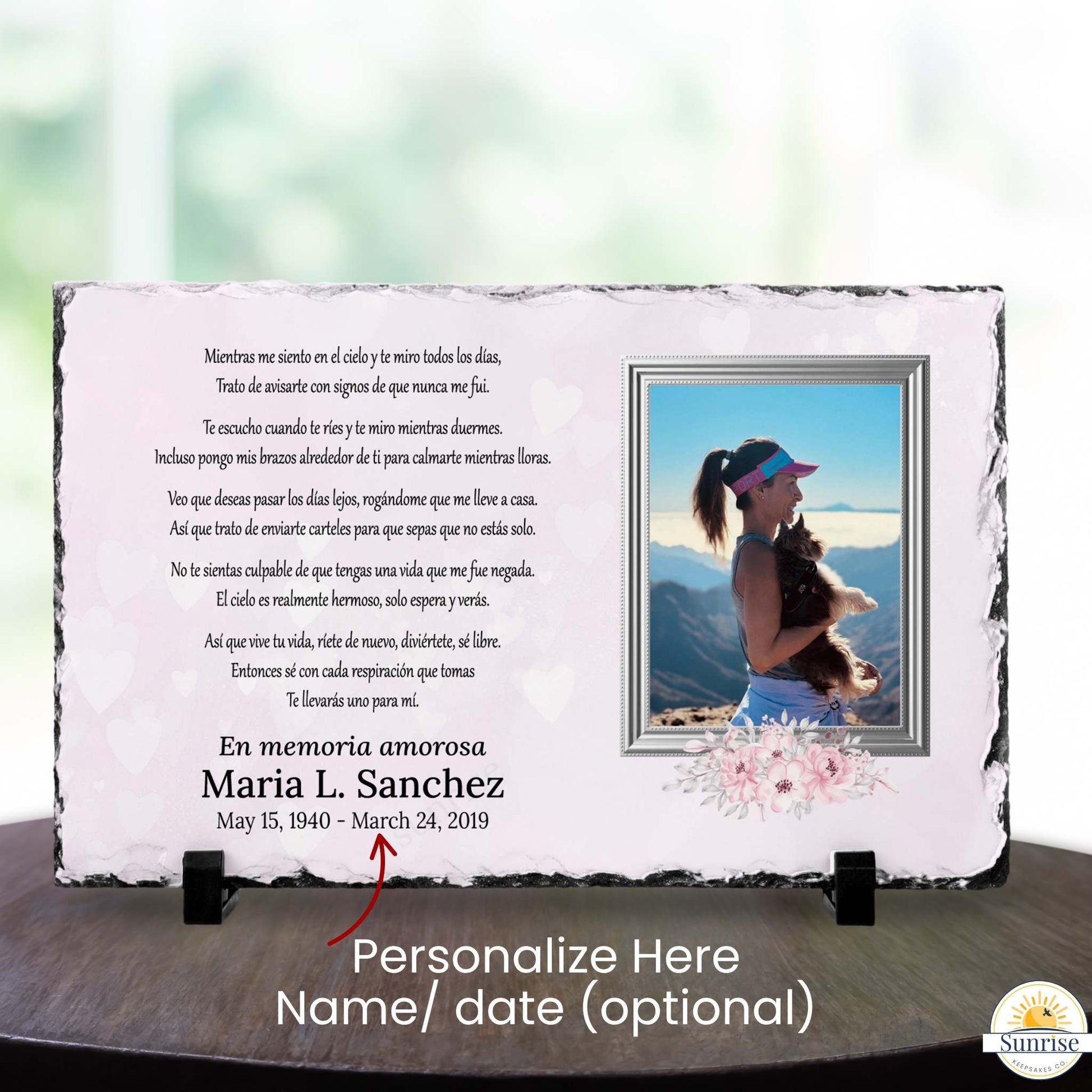 Personalized rectangular memorial stone plaque on a dark tabletop with a blurred light background. The plaque has a white center with black Spanish text, a photo of a woman with a dog, floral decoration, and the name 'Maria L.
