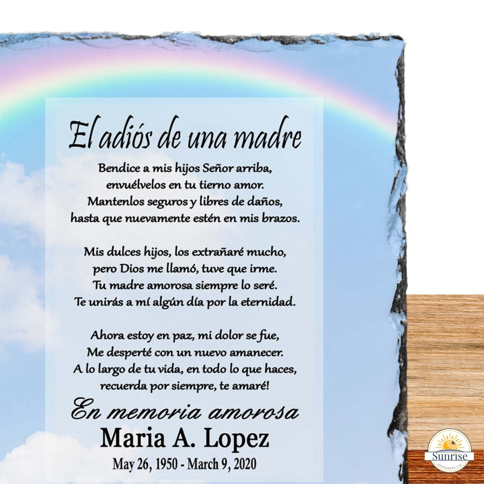 A memorial slate plaque with a printed Spanish 'adios de una madre' poem on a light blue sky background with a faint rainbow and clouds, shown leaning against a wood surface. 