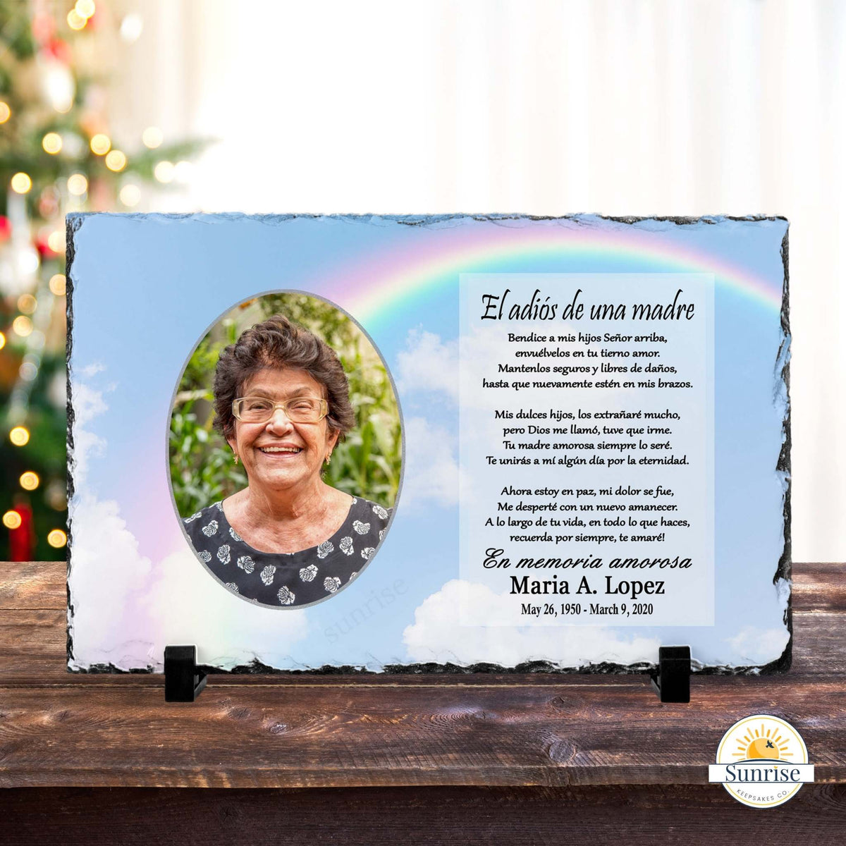 Personalized memorial slate plaque with a photo of a woman and Spanish text reading 'El adiós de una madre' and 'En memoria amorosa Maria A. Lopez', displayed on a wooden surface with blurred holiday lights in the background.
