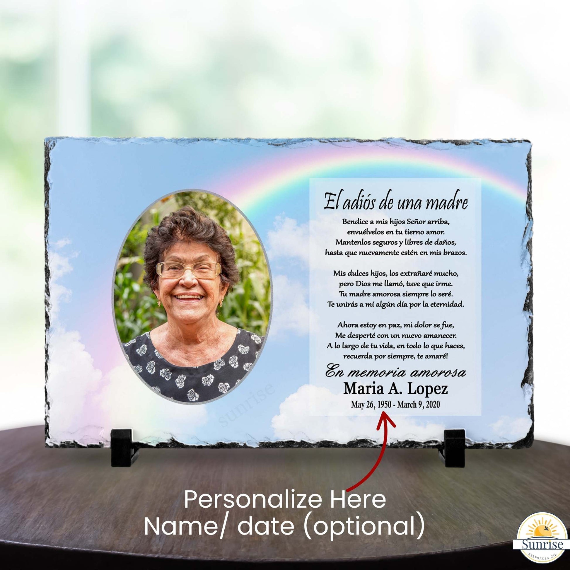 Personalized memorial slate plaque on a dark wooden surface with a blurred indoor background. The plaque has a blue sky and rainbow design, a photo of a woman, a Spanish remembrance poem, and the name 'Maria A.