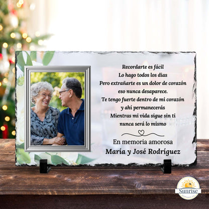 Personalized memorial slate plaque on a wooden surface with a blurred Christmas tree in the background. The white and green slate plaque features a photo of a couple and Spanish memorial text reading 'En memoria amorosa Maria y José Rodríguez'.
