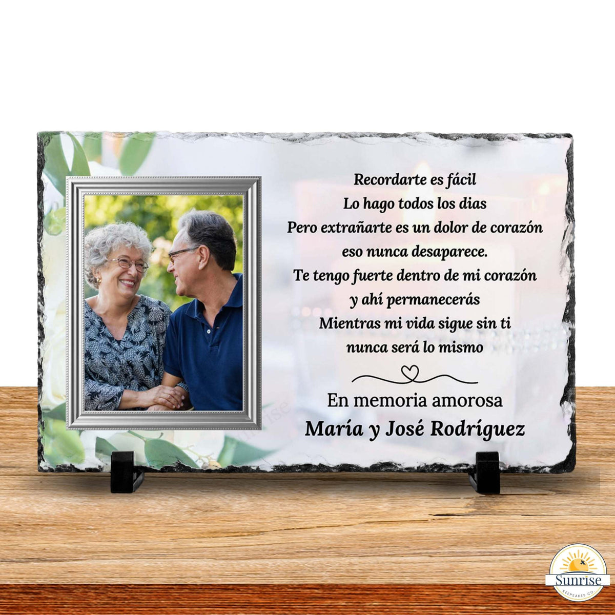 Personalized memorial slate plaque on a wooden surface with a photo of a couple and a Spanish remembrance inscription. The plaque has a white background with green leaf accents, black slate edges, and small black display stands.
