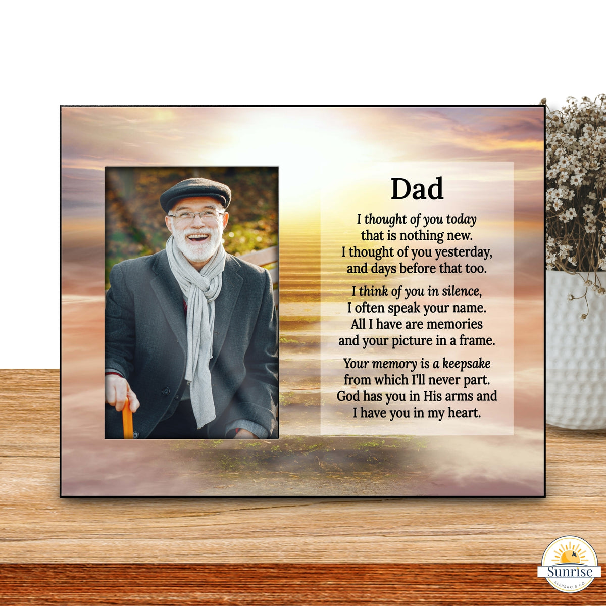 A square memorial plaque or photo frame displayed on a wooden surface, featuring a portrait of a smiling man on the left and a printed remembrance poem for 'Dad' on the right. The background shows a blurred indoor setting with a vase of flowers.